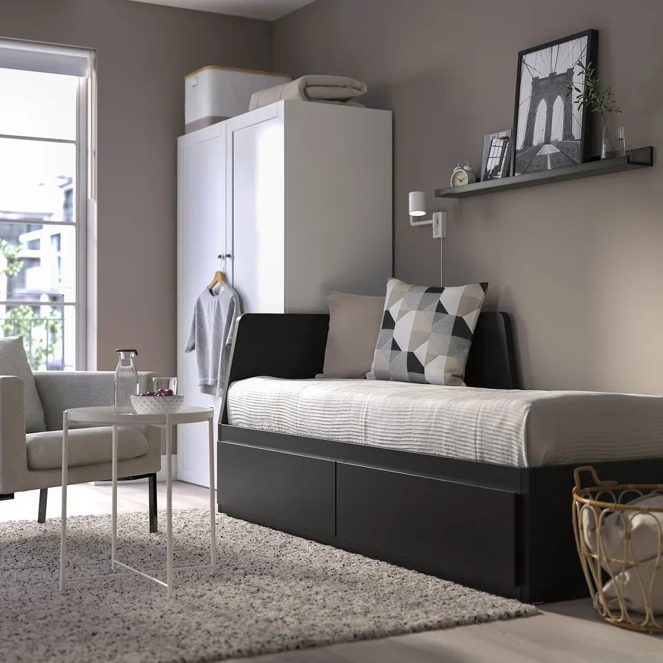 Modern grey bedroom with FLEKKE day-bed, white closet, & grey armchair. Day-bed has black drawers & geometric pillow.