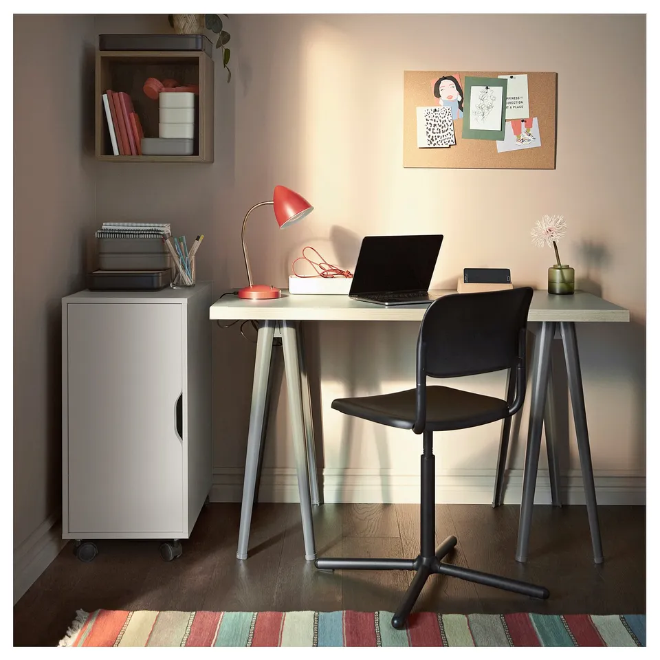 A home office setup featuring a white ALEX storage unit on wheels, a black office chair, and a desk with a laptop and red lamp.