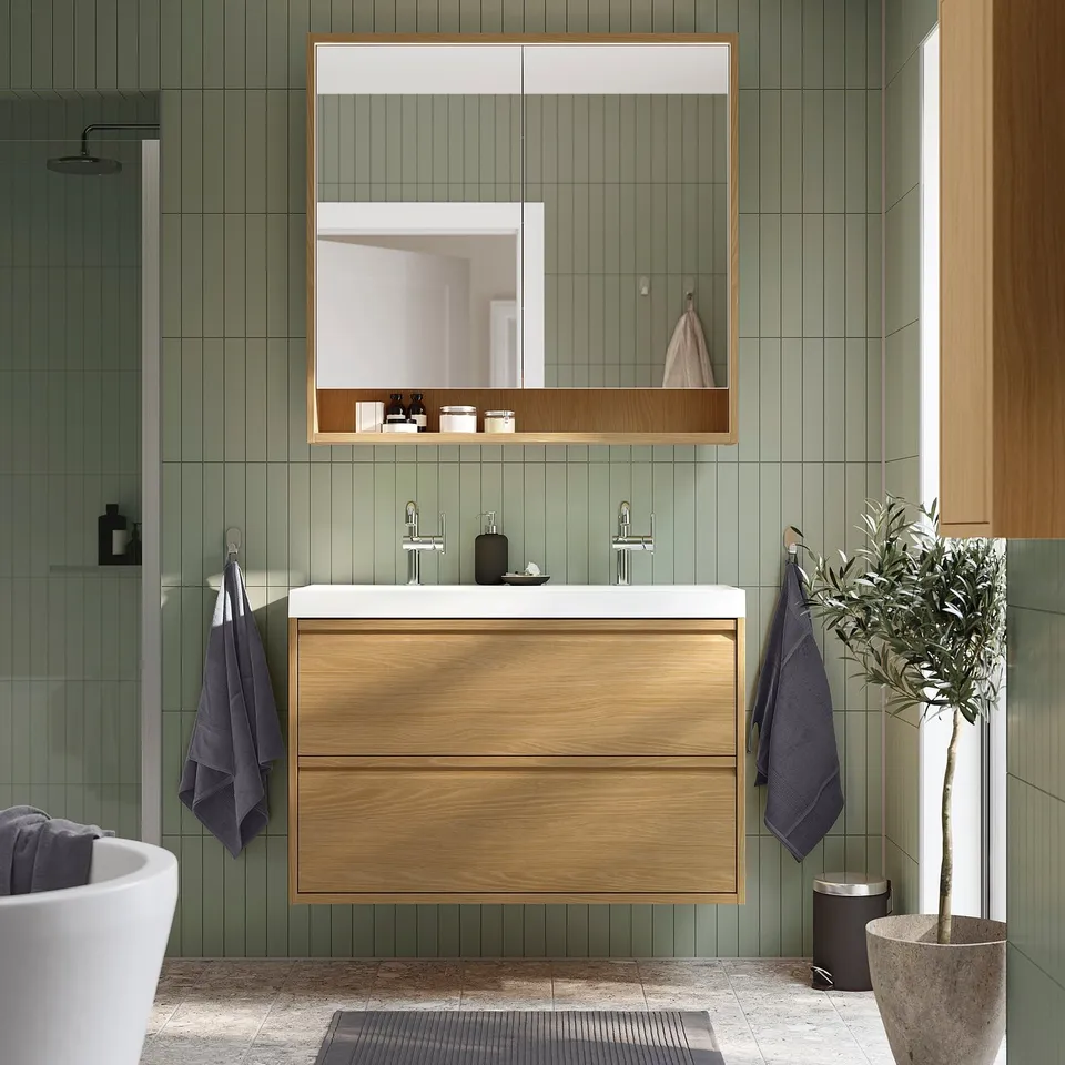 Modern green-tiled bathroom with wooden cabinetry. Wooden vanity, mirror, and dark grey towels. Potted plant to the right. Contemporary and relaxing.