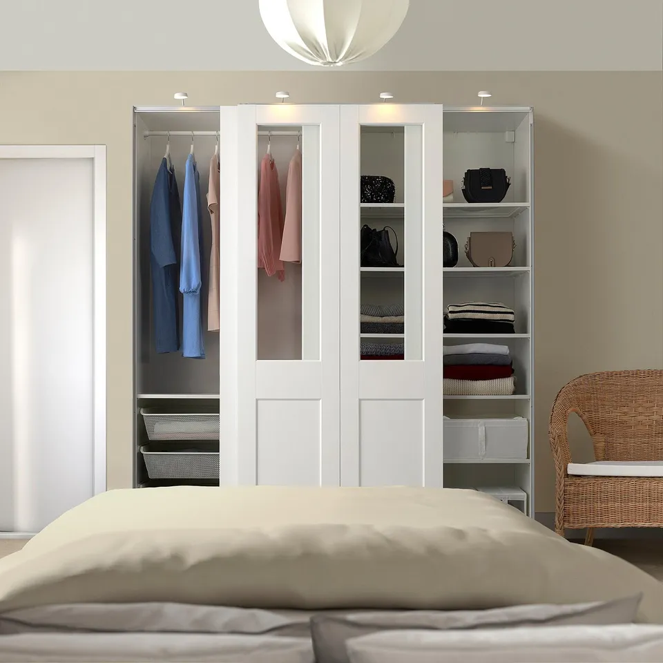 This image a white PAX wardrobe displaying clothes, bags, and folded clothes, with a bed in front.