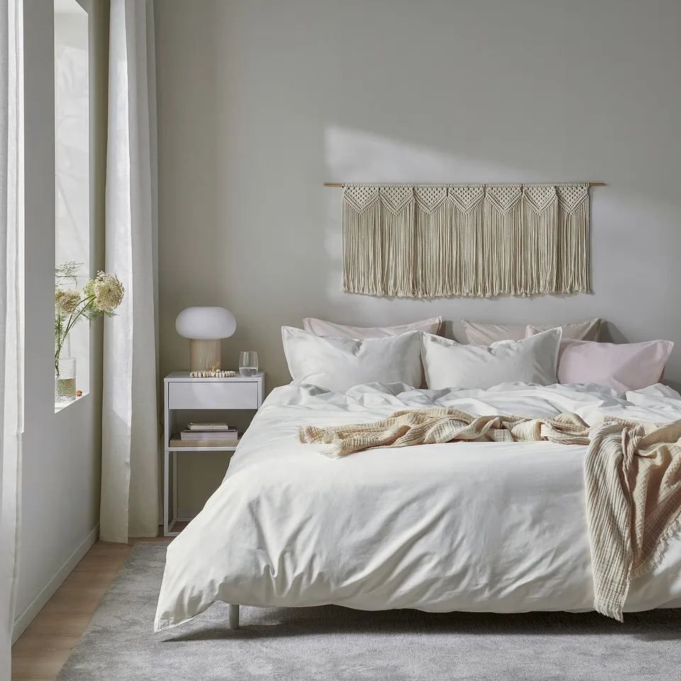 Minimalist bedroom with ÄNGSLILJA duvet, throw, bedside lamp, flowers, macrame, and natural light.