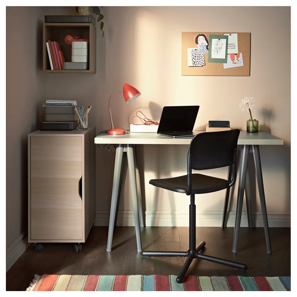 A modern home office with wooden storage units, a light wooden desk, black chair, red lamp, and a corkboard with various notes and images.