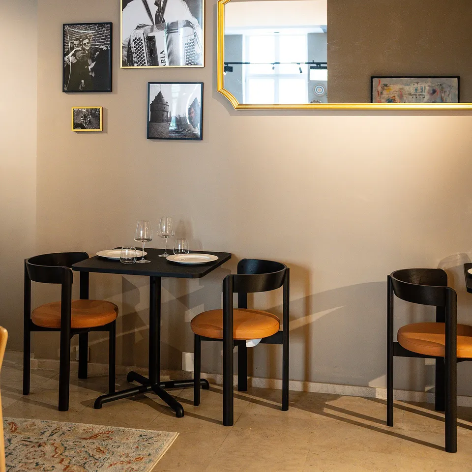 Modern dining setup with black table, four leather chairs, wine glasses, plates. Art and mirror on wall, rug below table.