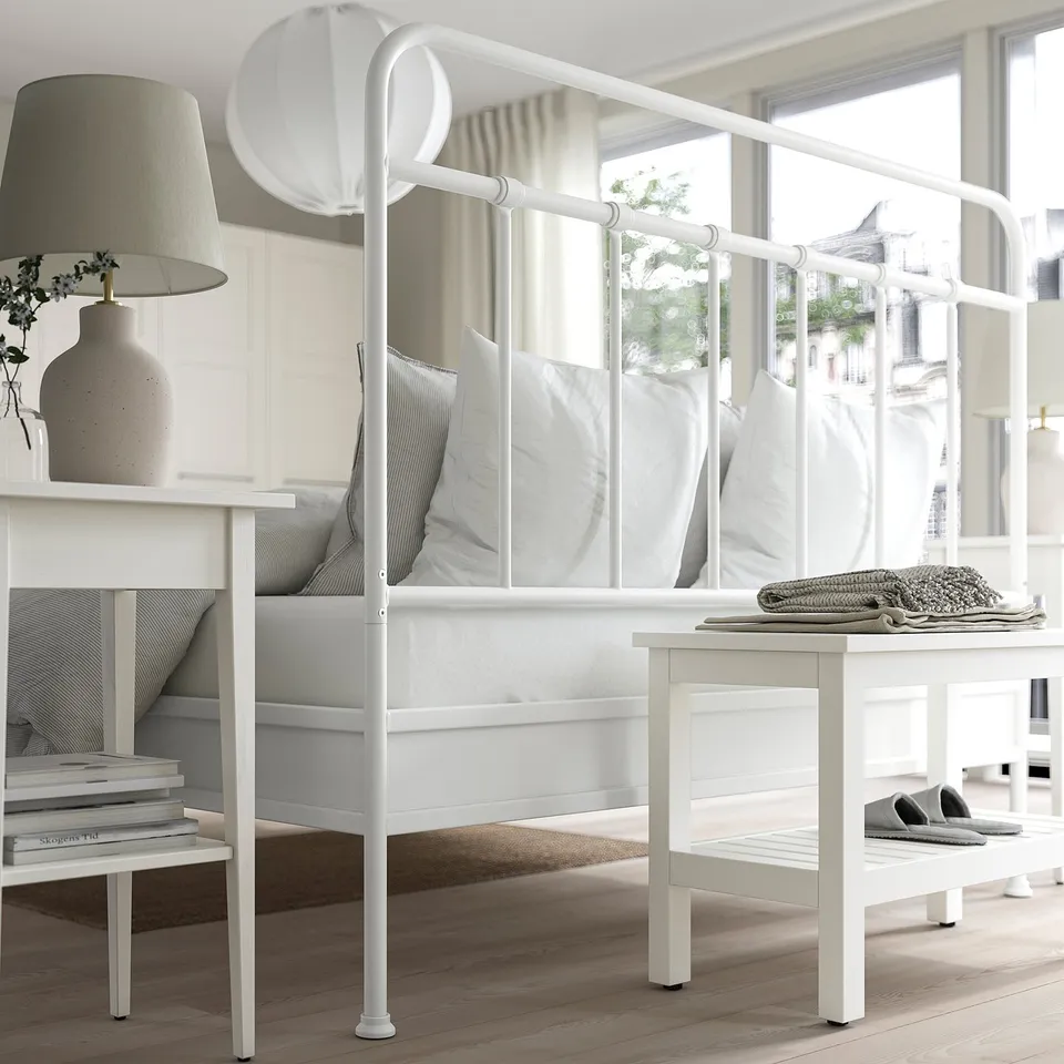 Modern white metal bed with curved headboard, cosy blankets, next to a bedside table with lamp. Soft pillows, large window showing city view.