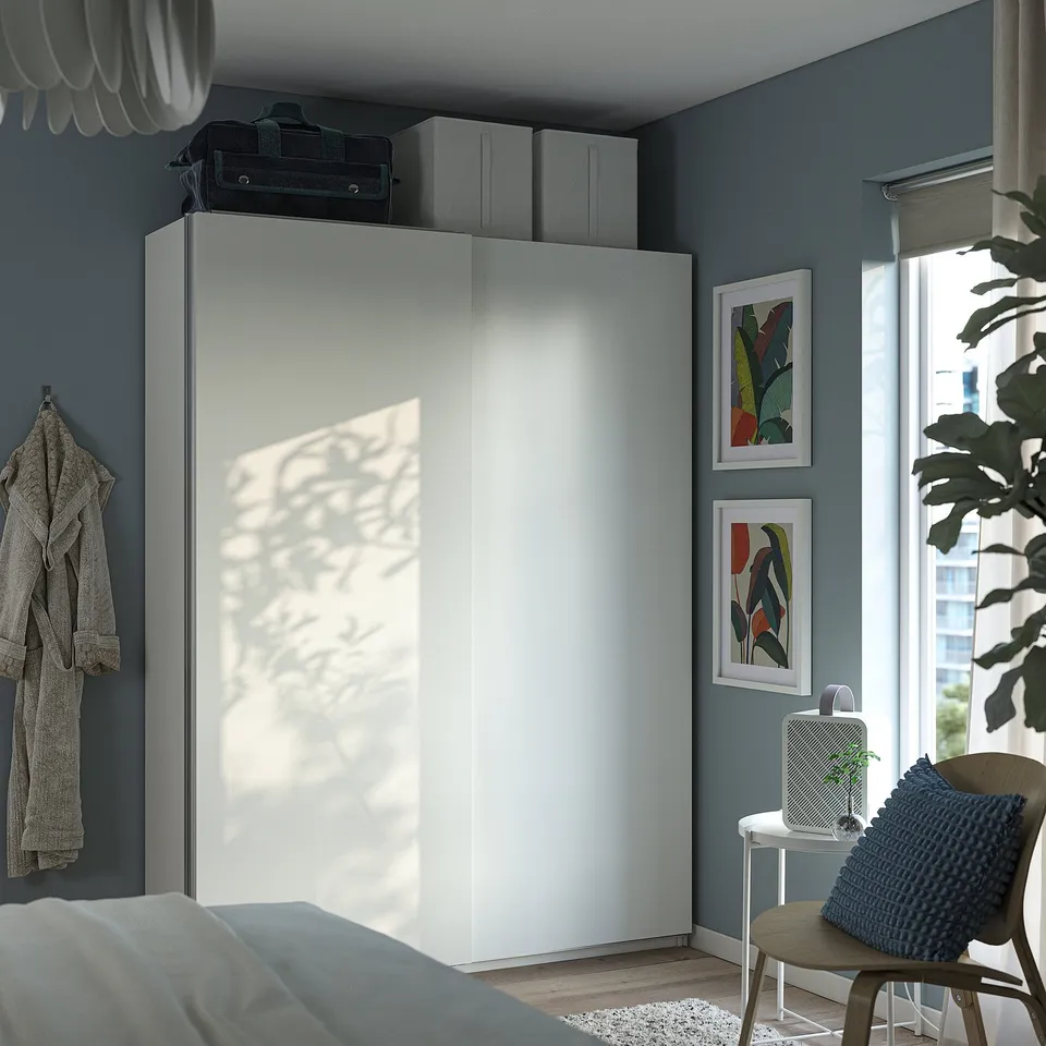 Modern blue bedroom with large white PAX wardrobe, sliding doors, and grey wall hooks. Decorated with colourful art and a cosy armchair.