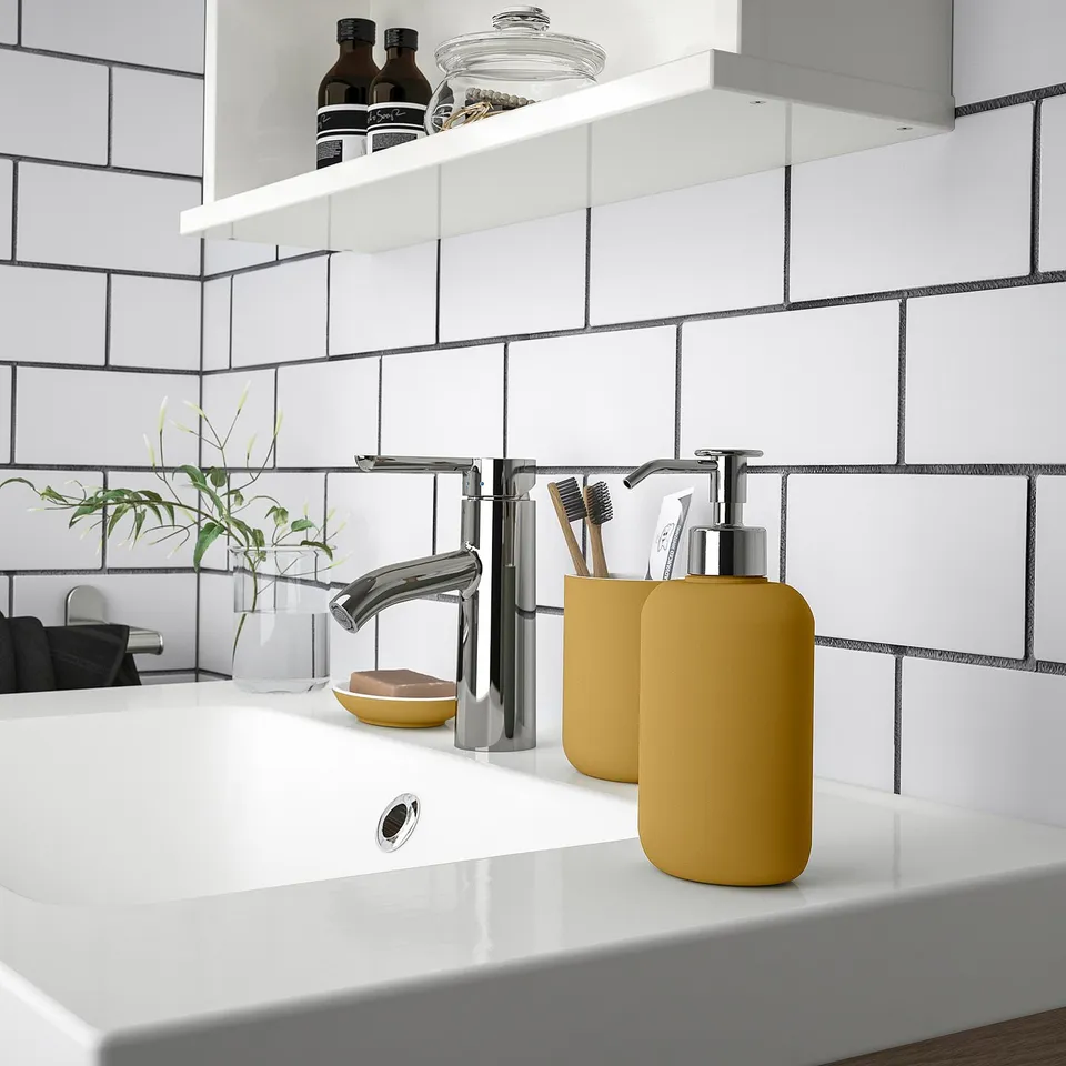 Modern bathroom sink with chrome tap, yellow soap dispenser, and accessories neatly arranged on a white countertop and tiled wall.