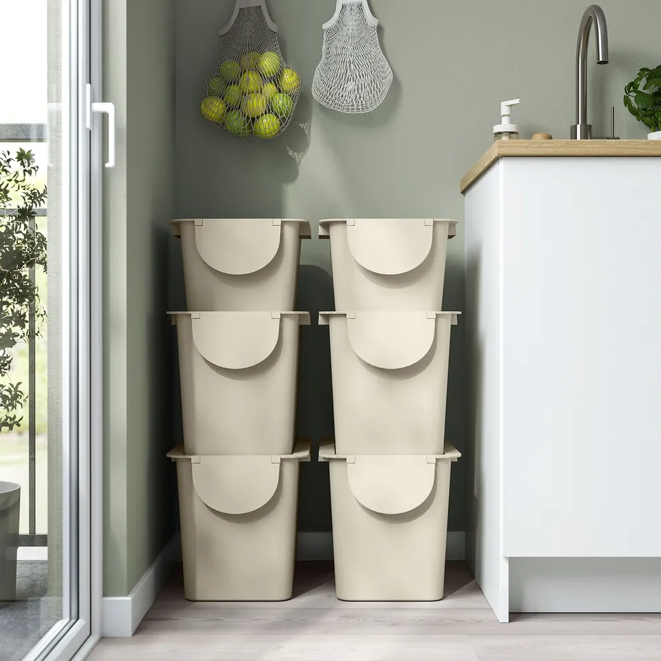 Two stacked waste bins with lids in a kitchen. Hanging net bags with fruits nearby.