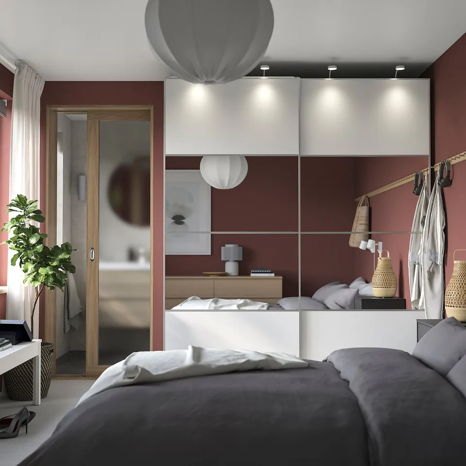 This modern bedroom a stylish mirrored & white sliding closet door, grey bedding, a cosy plant, & a sleek bathroom entrance.