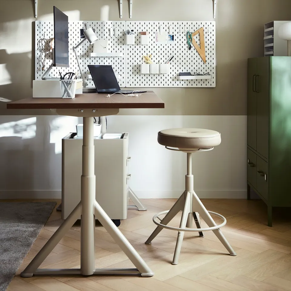 Minimalist home office: white metal desk with wooden top, swivel stool, pegboard, laptop, and accessories.