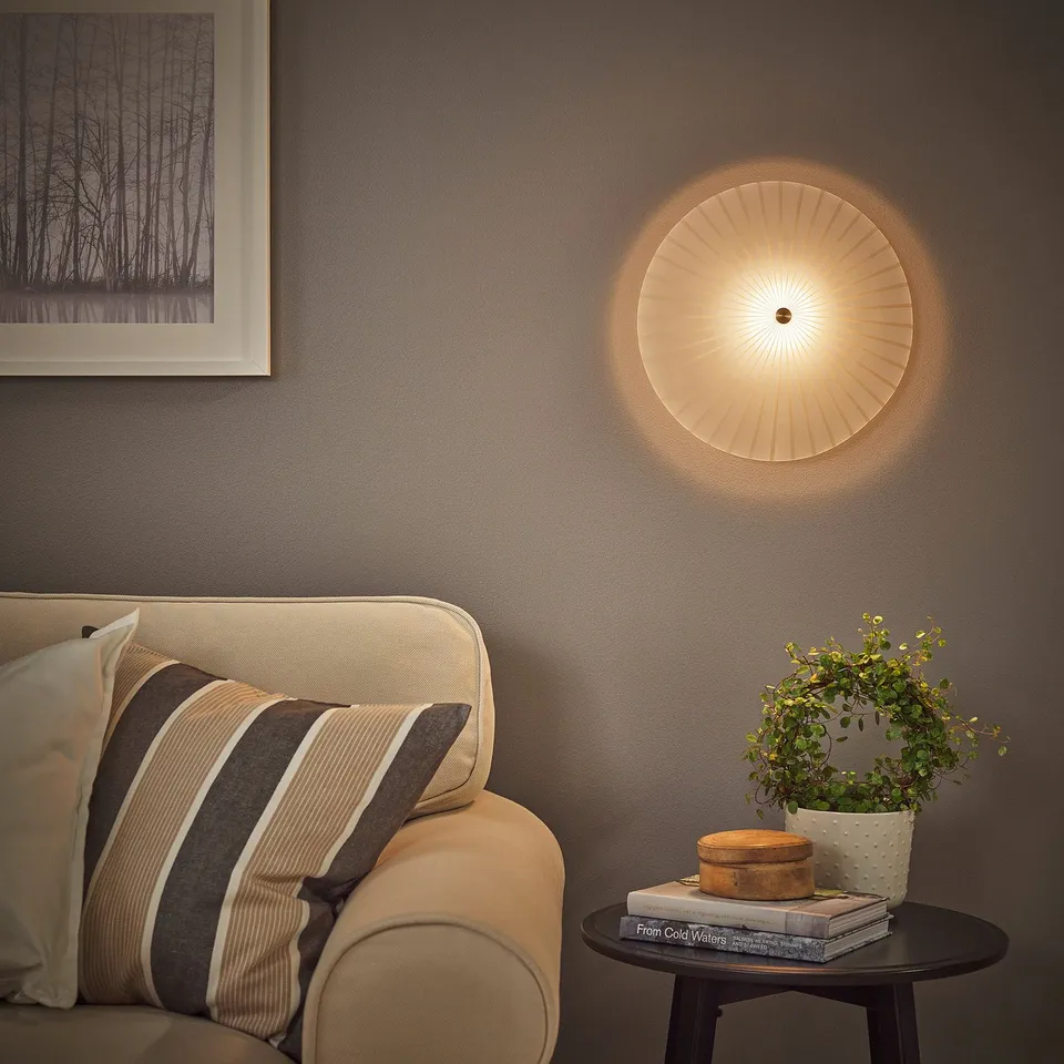 Modern living room with RORKULT wall lamp above a beige sofa, coffee table with plant, books and decor. Dark grey wall contrasts with lamps warm glow.