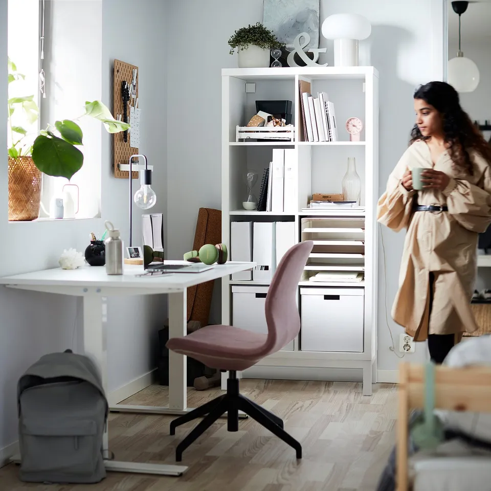 A person smiles, holding a cup, in a well-lit room with a white KALLAX shelving unit filled with books, decorative items, and plants.