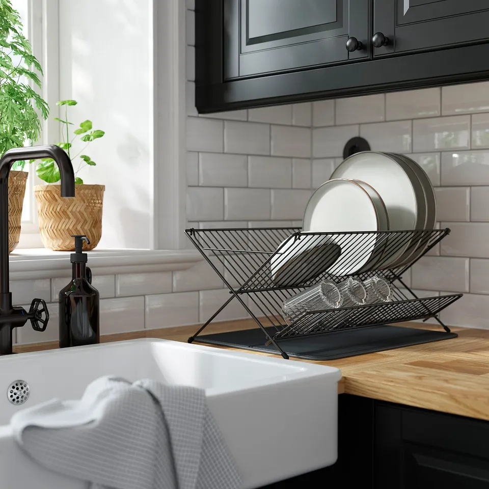 Kitchen counter with white sink, black tap, and NYSKÖLJD mat holding clean dishes. Wood countertop, grey backsplash, and potted plant nearby.