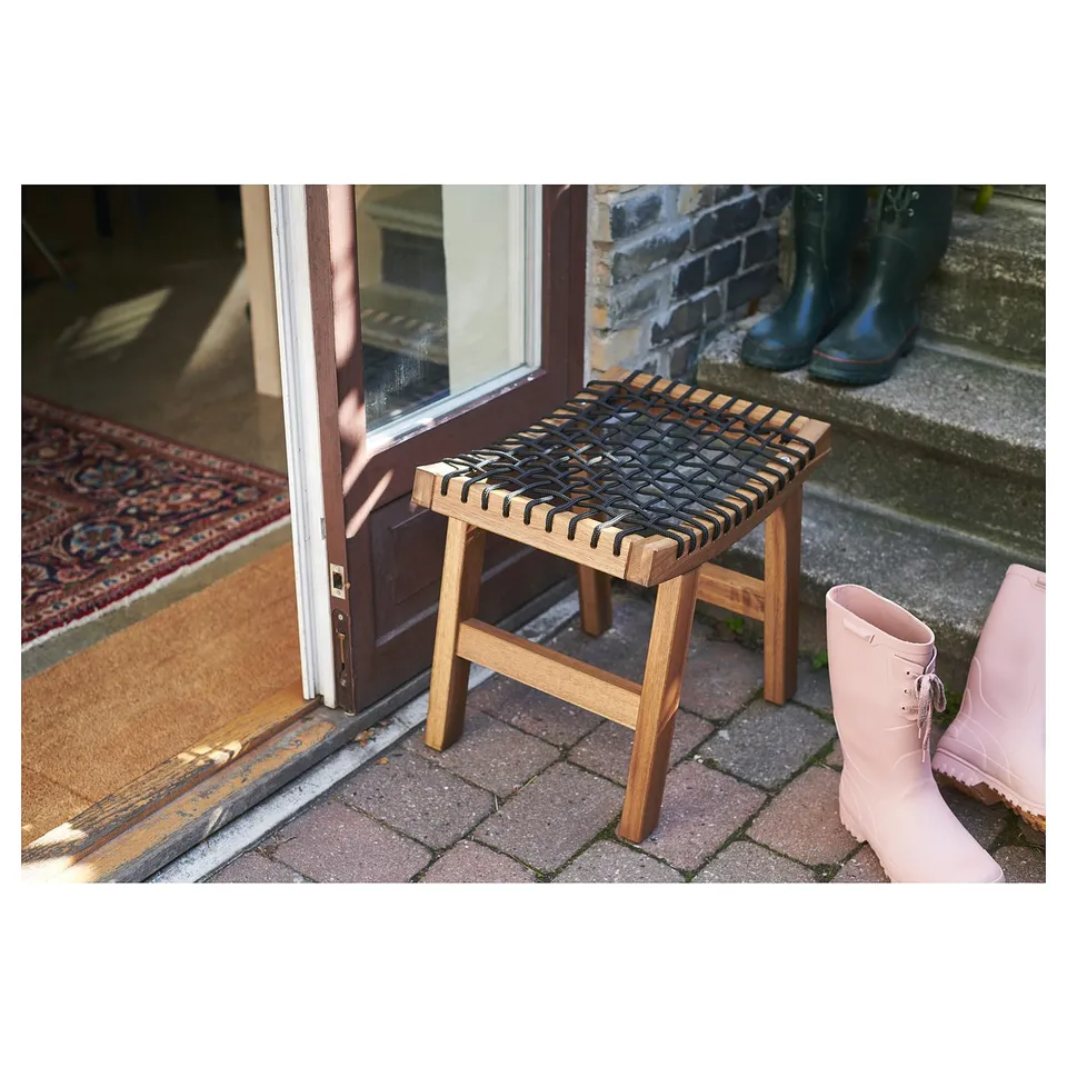 Outdoor stool named STACKHOLMEN, made of acacia wood. Brown, woven seat, four legs, placed near an entrance door.