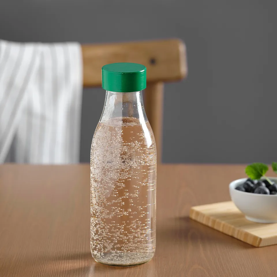 Clear glass bottle with green lid, standing on wooden table, designed for reusing beverages, fits in backpack or car holder.
