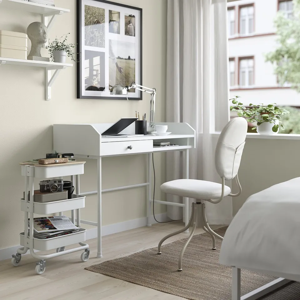 White RÅSKOG trolley under desk, versatile storage, near chair and window.