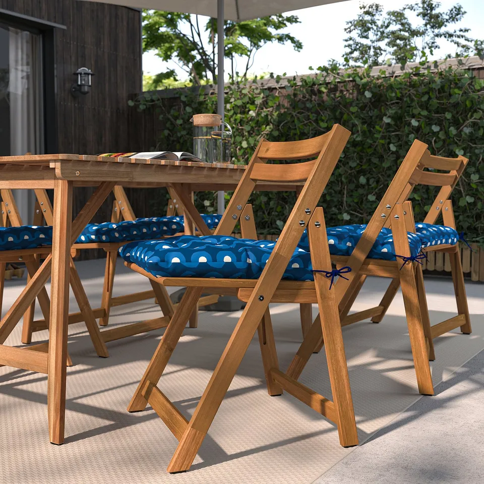 Four KLÖSAN chairs with blue cushions around a table, creating a cosy patio seating area with greenery.