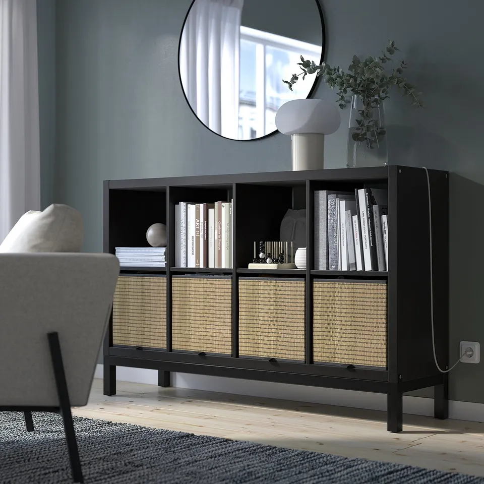 Modern living room with black KALLAX shelf, housing books, vases, and plants. Miror above, chair to the side, white rug underneath.