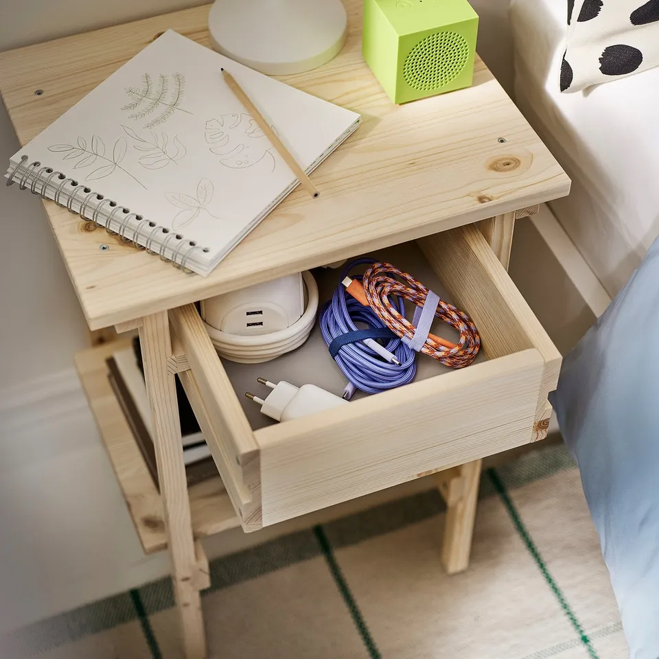 Wooden bedside table with open drawer showing cables, charger, and notebook; green speaker and lamp on top.