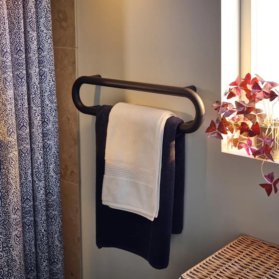 Dark grey BÄSINGEN towel rail with white towel, rounded edges. Decorative plant beside it.