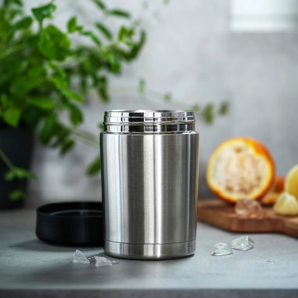 Silver insulated food container on a grey countertop, surrounded by ice, lemon halves, and a plant.