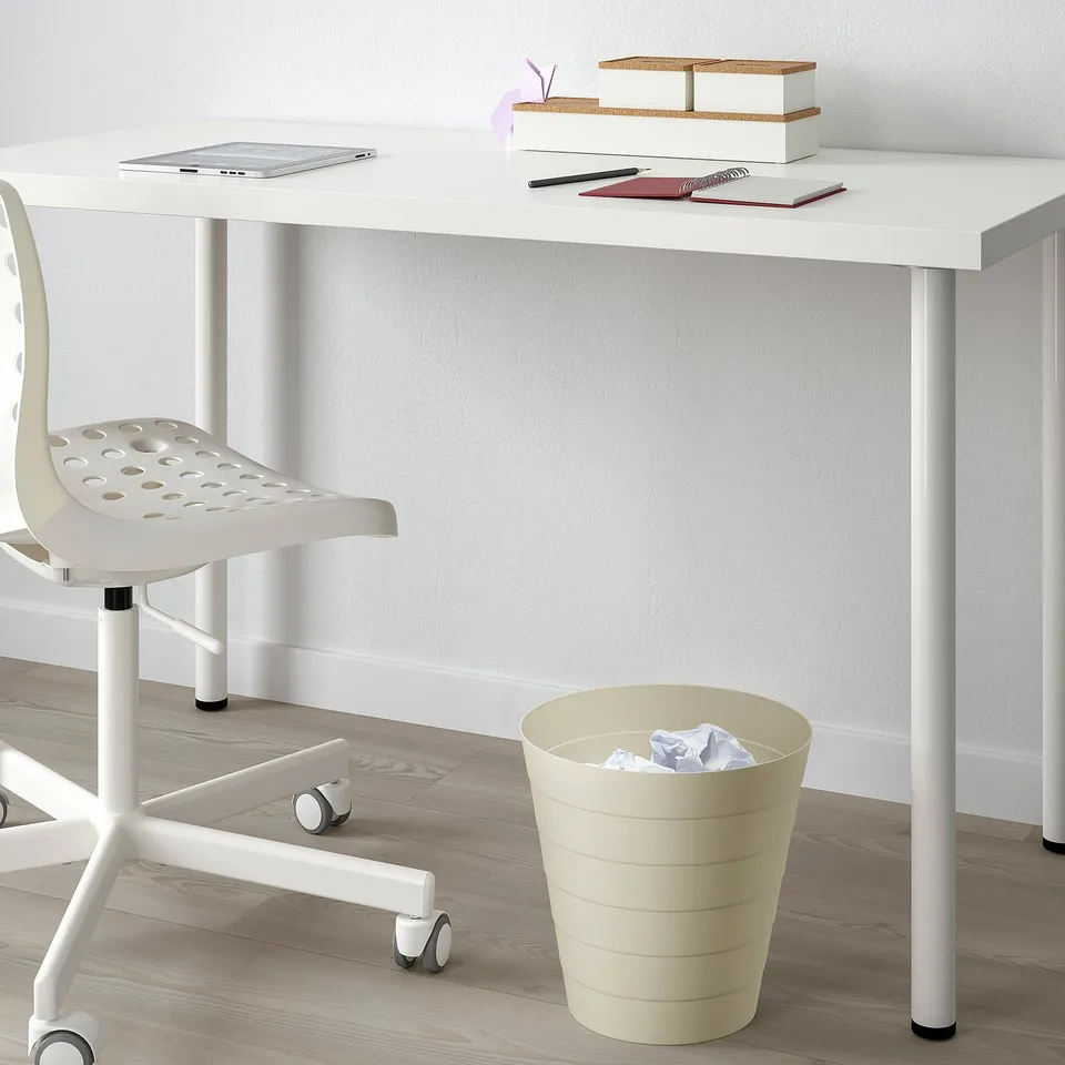 A minimalist workspace with a white desk, a white chair, and a beige waste bin holding paper. The desk has a laptop, notebooks, and storage boxes.