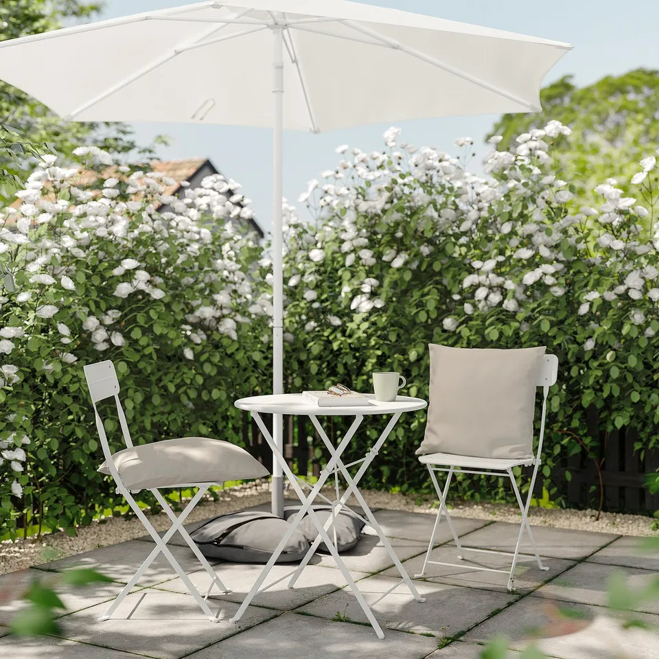 Peaceful patio setting with white HJÄRTERÖ cushions on chairs, under a white umbrella, surrounded by blooming flowers.