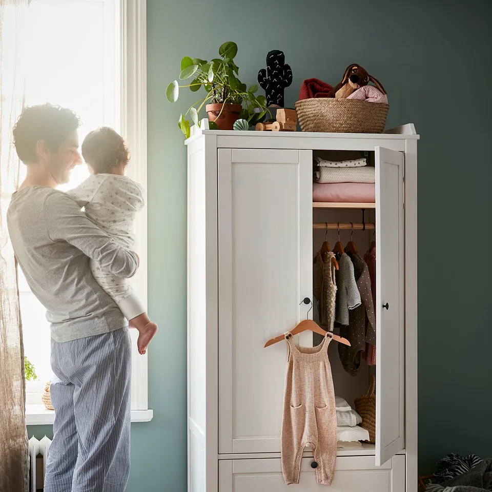 Person holding child near open white wooden cabinet with clothes; hanging garment, folded clothes, and a wicker basket on top.