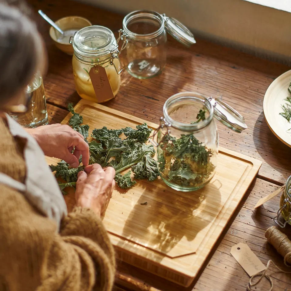 Person preparing leafy food on a wooden board, using KORKEN jars for storage, which have airtight seals to preserve freshness.