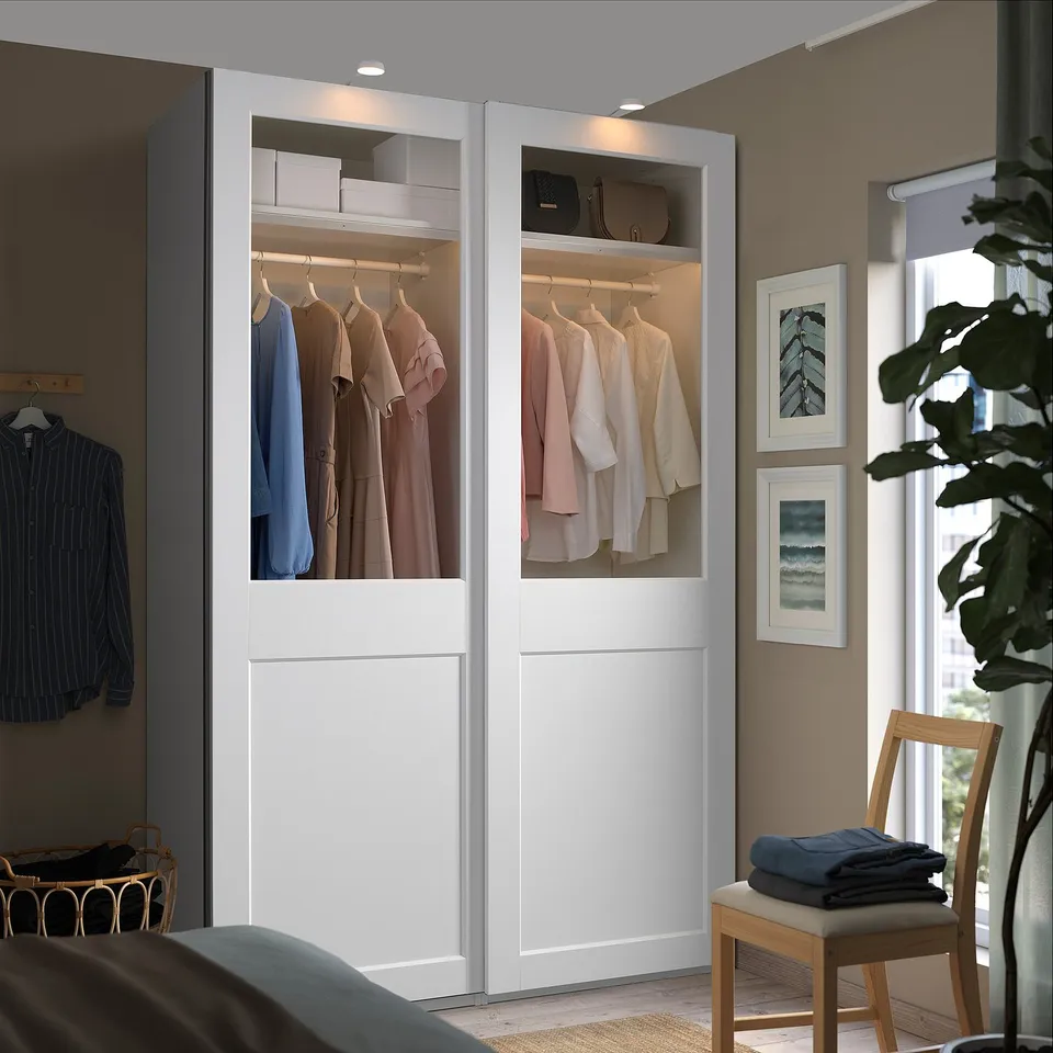 Modern wardrobe with glass doors displays clothes, blending with beige walls and large windows. A chair holds folded clothes next to it.