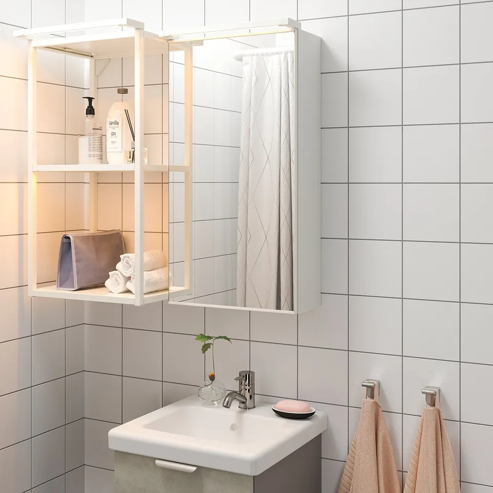 Modern bathroom with white tiles. Led-lit cabinet, mirrored doors, and open shelves with toiletries and towels. Peach towels on hooks below sink.