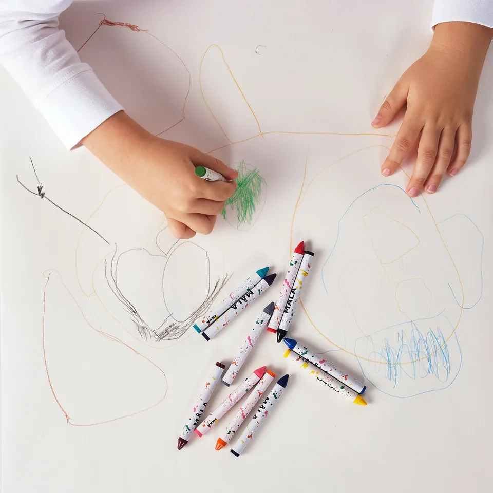 Childs hands drawing with colourful crayons. Several MÅLA crayons are scattered on the paper, which abstract drawings in various colours.
