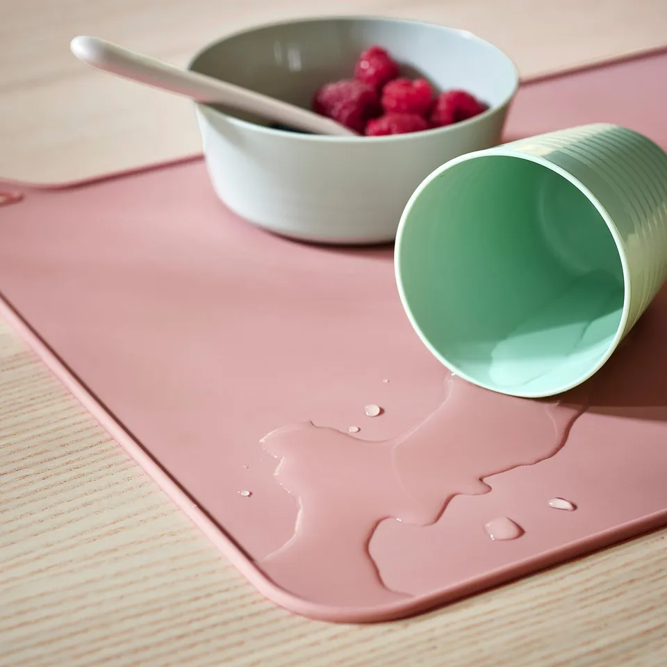 Silicon place mat with cat design, holding bowl of berries and tipped cup, protects table from spills and scratches.