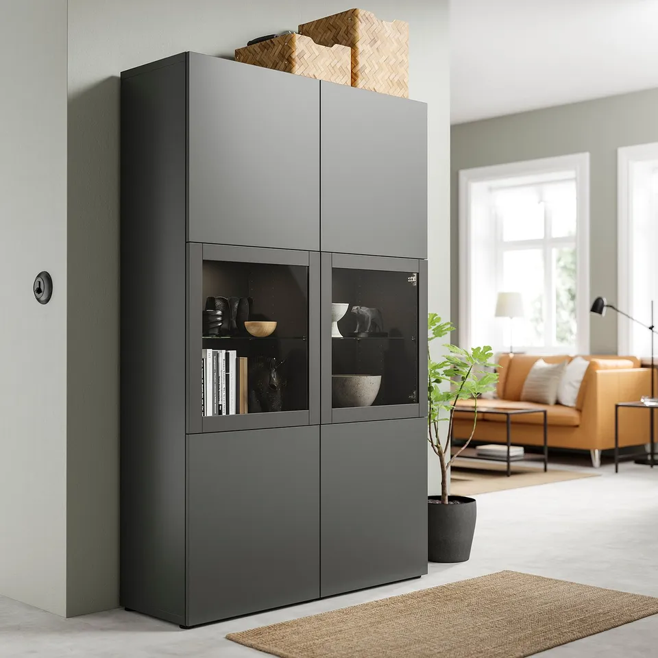 Dark grey BESTÅ storage unit with glass doors; modern, rectangular design; holds decorative items, books, and a woven basket.