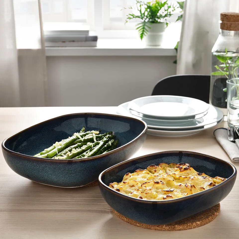 Two blue oven-safe dishes on a table hold asparagus with cheese and a baked dish, near stacked plates and a plant.