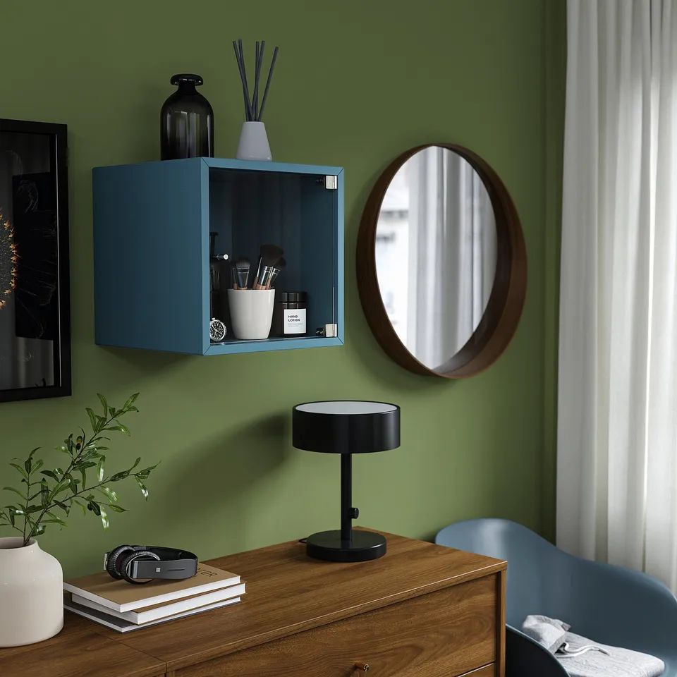 Teal EKET shelf with glass doors storing makeup, next to wooden mirror and lamp on dresser, green wall.