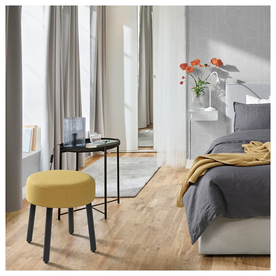 Bright, yellow MÖRTFORS stool with black legs sits next to a grey bed with dark grey covers and a wooden table with a vase of orange flowers.