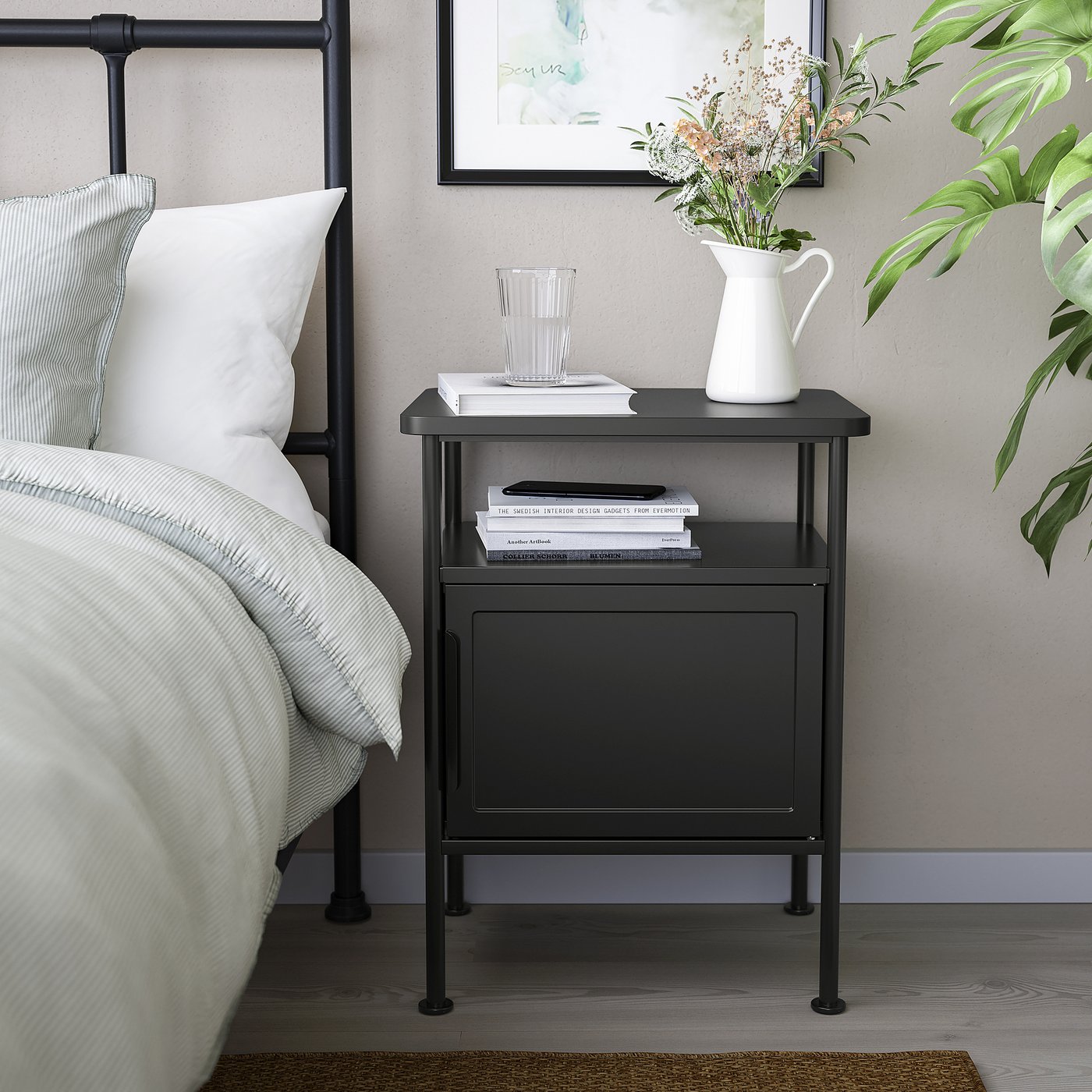 Modern bedroom setup. Small black bedside table next to bed holds books, glass, and flowers. Large bed with white linens and striped pillows at left.
