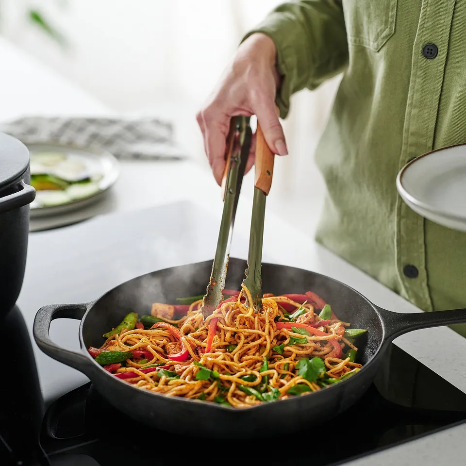 A person uses kitchen tweezers to cook noodles in a pan. The tweezers have wooden handles for a good grip.