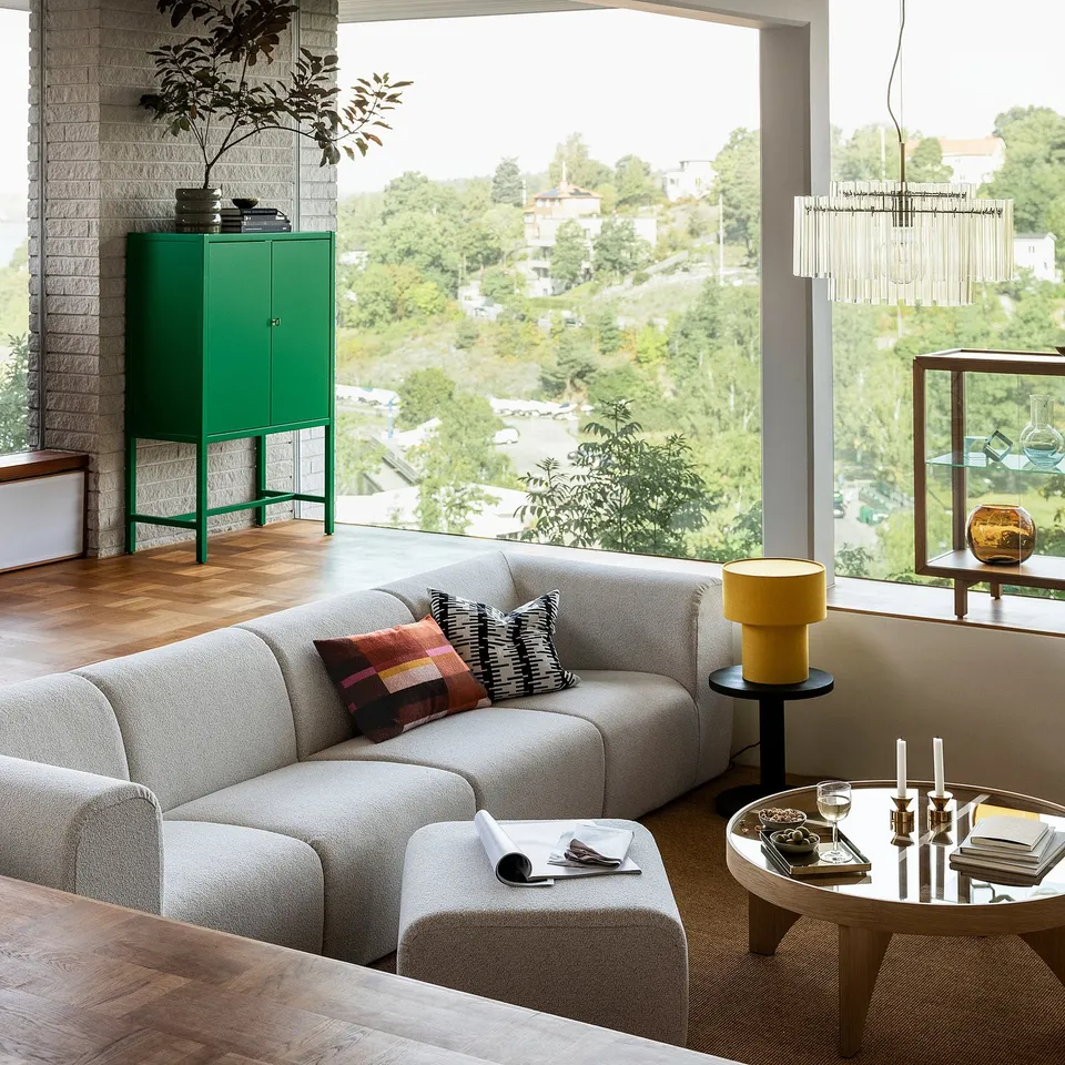 Modern living room with large grey sectional sofa, colourful pillows, wooden coffee table, green cabinet, yellow lamp, and large window showing trees.