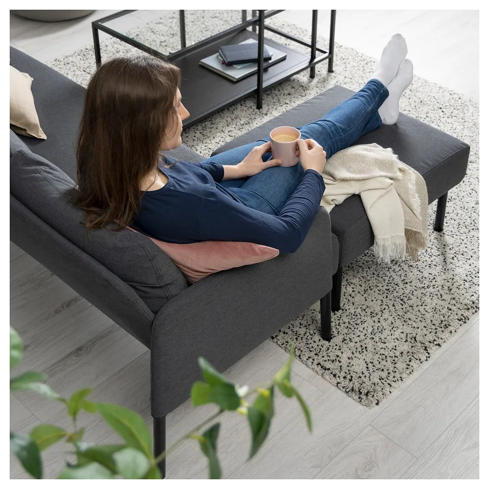 A person relaxes on a grey GLOSTAD sofa, holding a pink mug, with feet up, enjoying a tranquil moment in a modern living space.