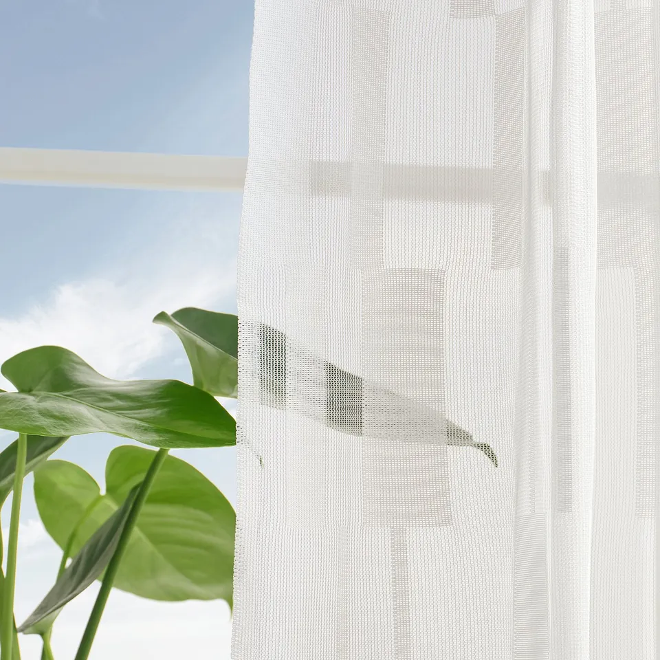 Window with sheer curtains, pattern, and multi-header tape, showing leafy green outside view.
