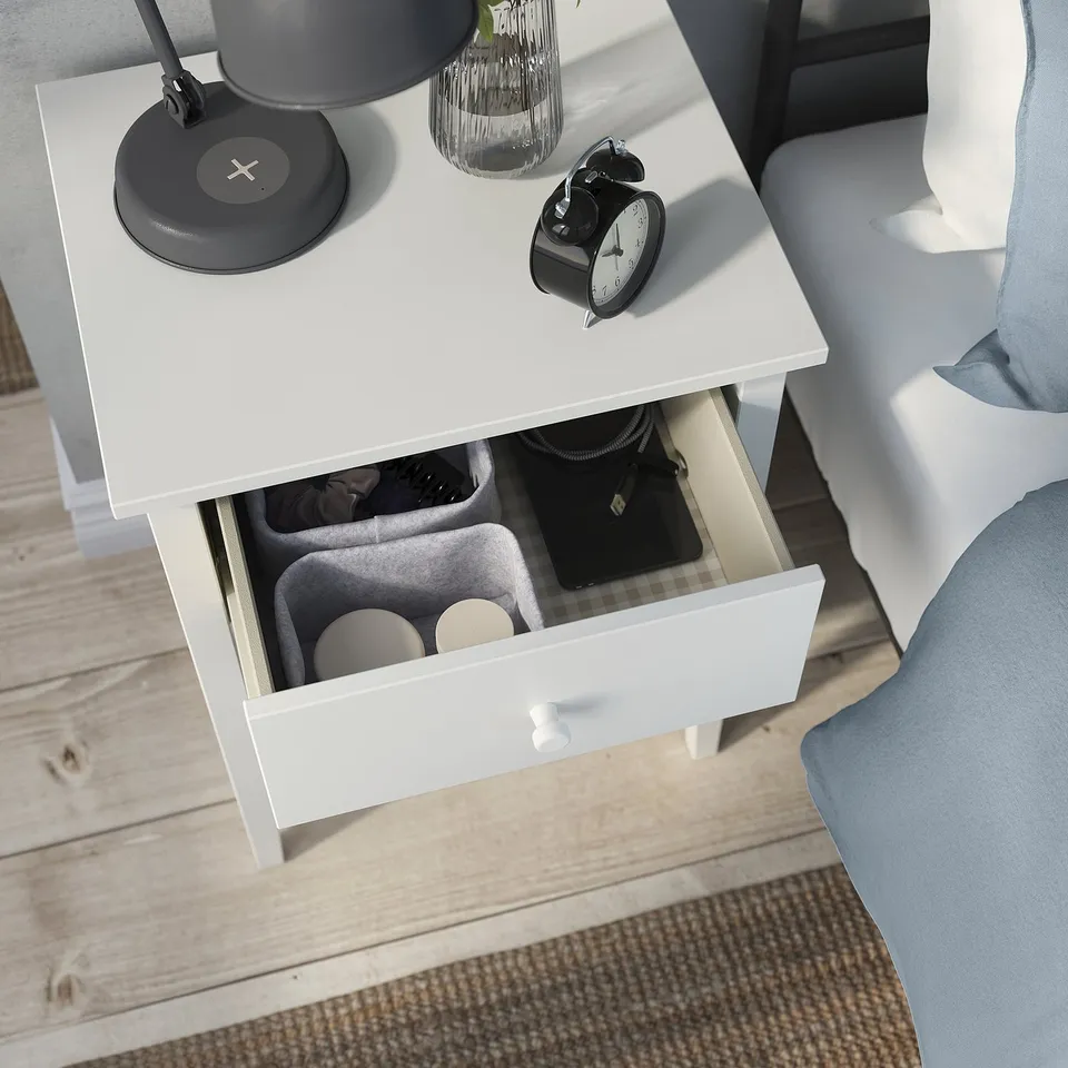 White bedside table with open drawer revealing organised items: headphones, wallet, and sunglasses. Top has a lamp, vase, and alarm clock.