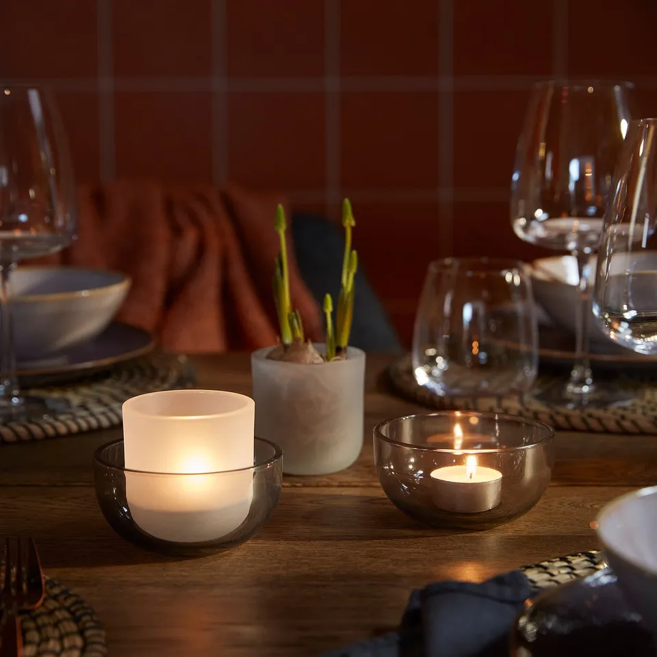 Wooden table set for dining, featuring lit tealight holders casting warm light, surrounded by glasses, plates, cutlery, and a small potted plant.