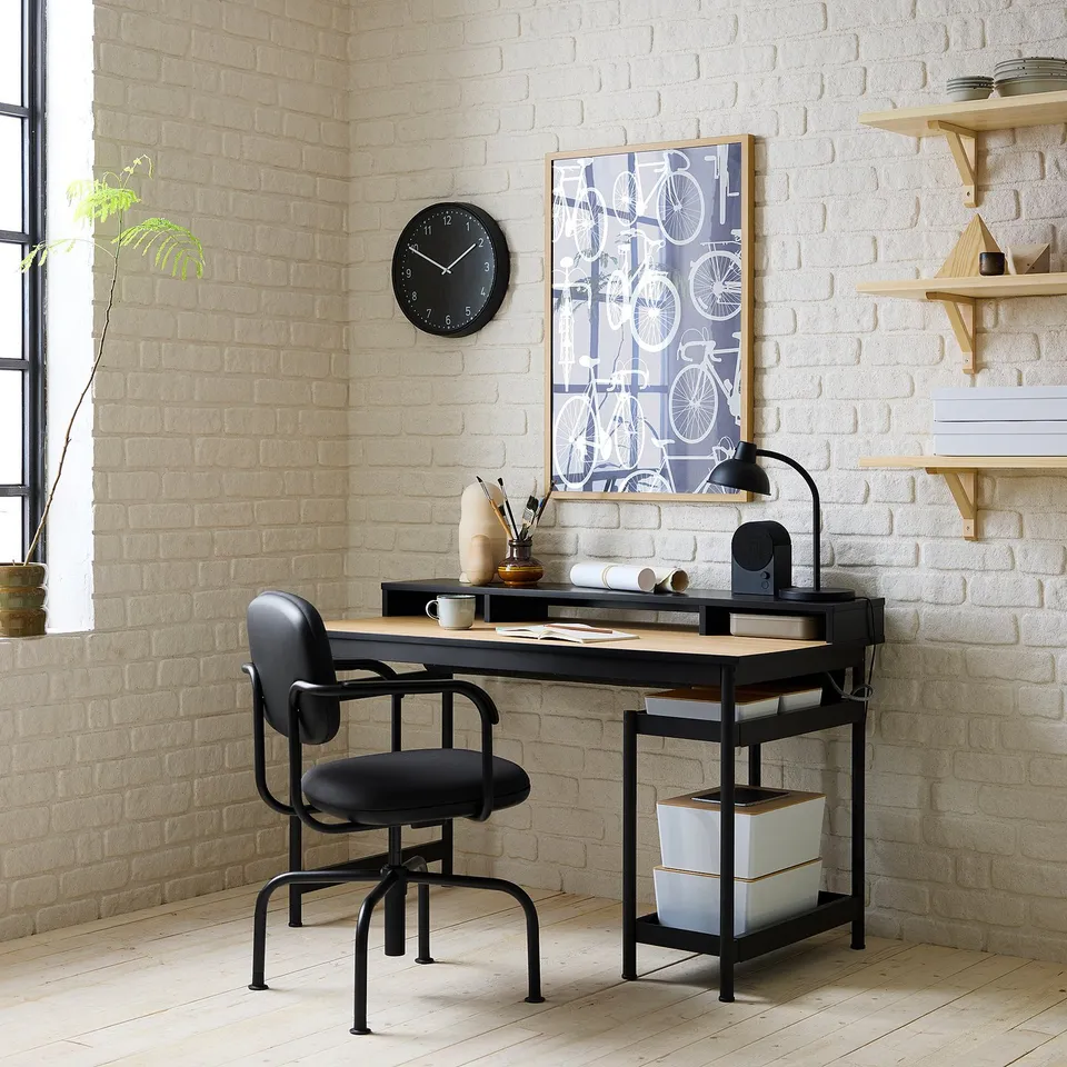 Modern black desk & adjustable chair against white brick wall with clock & art.