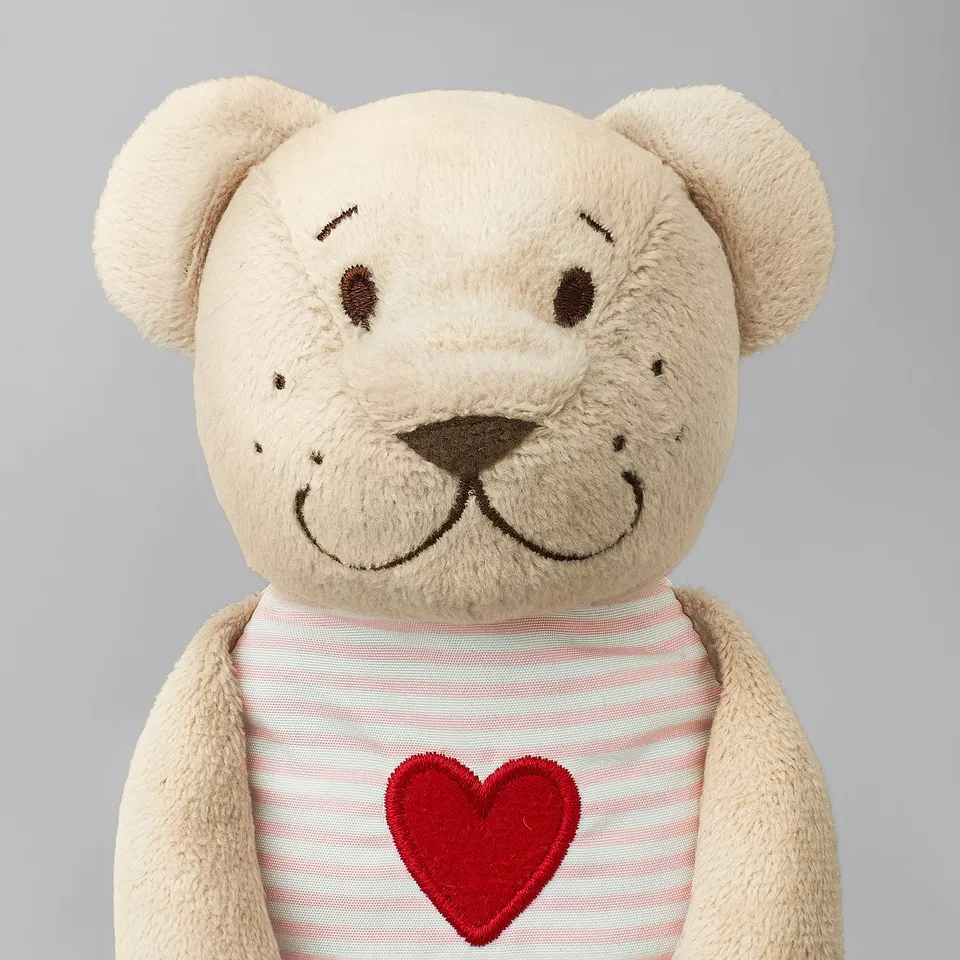A beige teddy bear with a red heart-shirt, soft and plush, smiling. Ideal for cuddling and play.