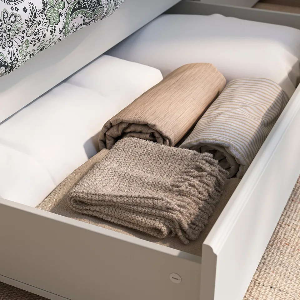 Open white bed drawer with neatly folded blankets: grey knit, beige striped, and waffle-weave.