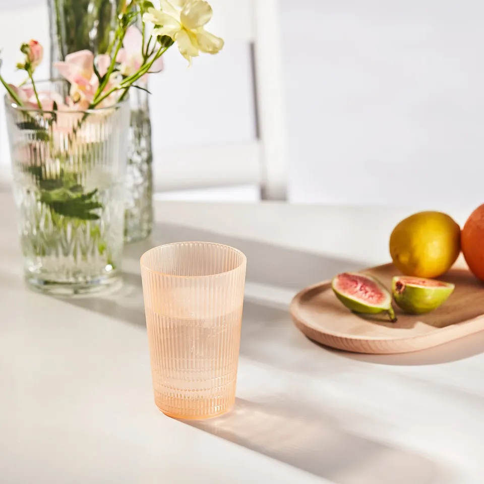 A peach-colored plastic glass sits on a table beside a plate of sliced fruits and a vase with flowers.
