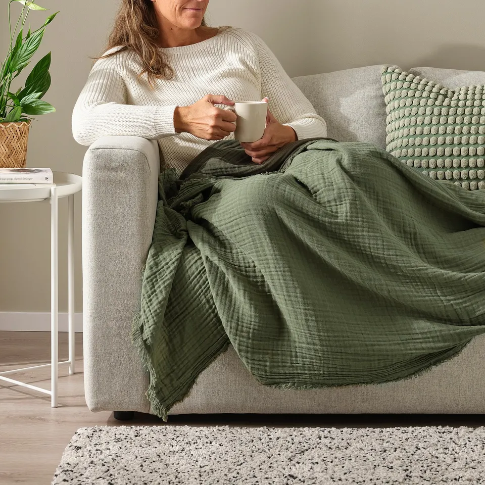 Person sitting on a beige couch, holding a white mug, with a green throw blanket over their lap.