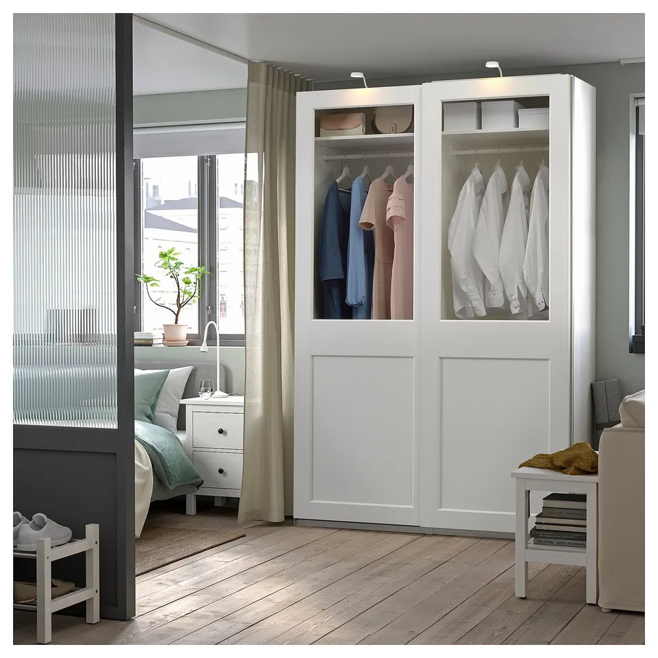 Modern bedroom with white PAX wardrobe, clear glass doors, hanging clothes, shelves, and built-in lights.