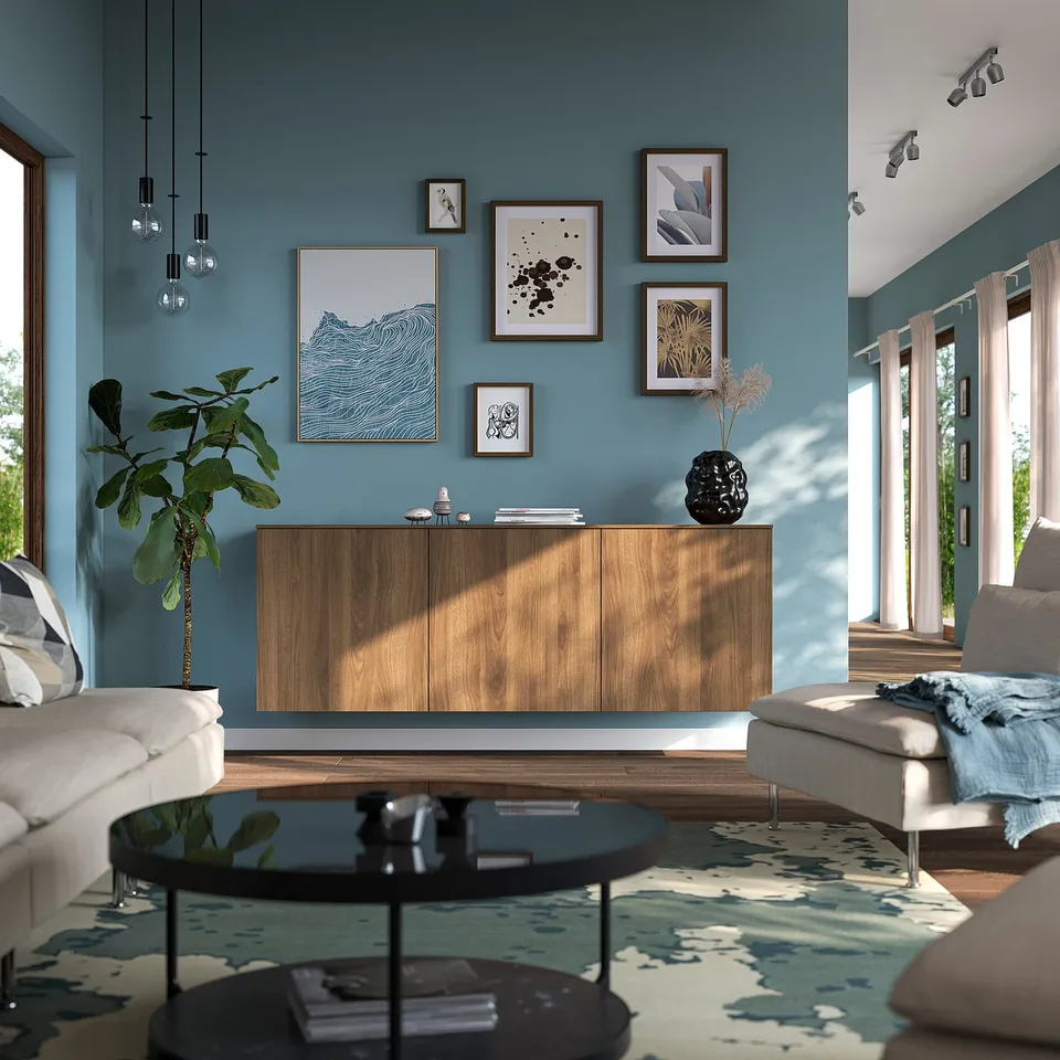 Modern living room with teal wall, BESTÅ sideboard, white sofa, ottoman, and glass table. Decorated with art, plant, rug, and lights.